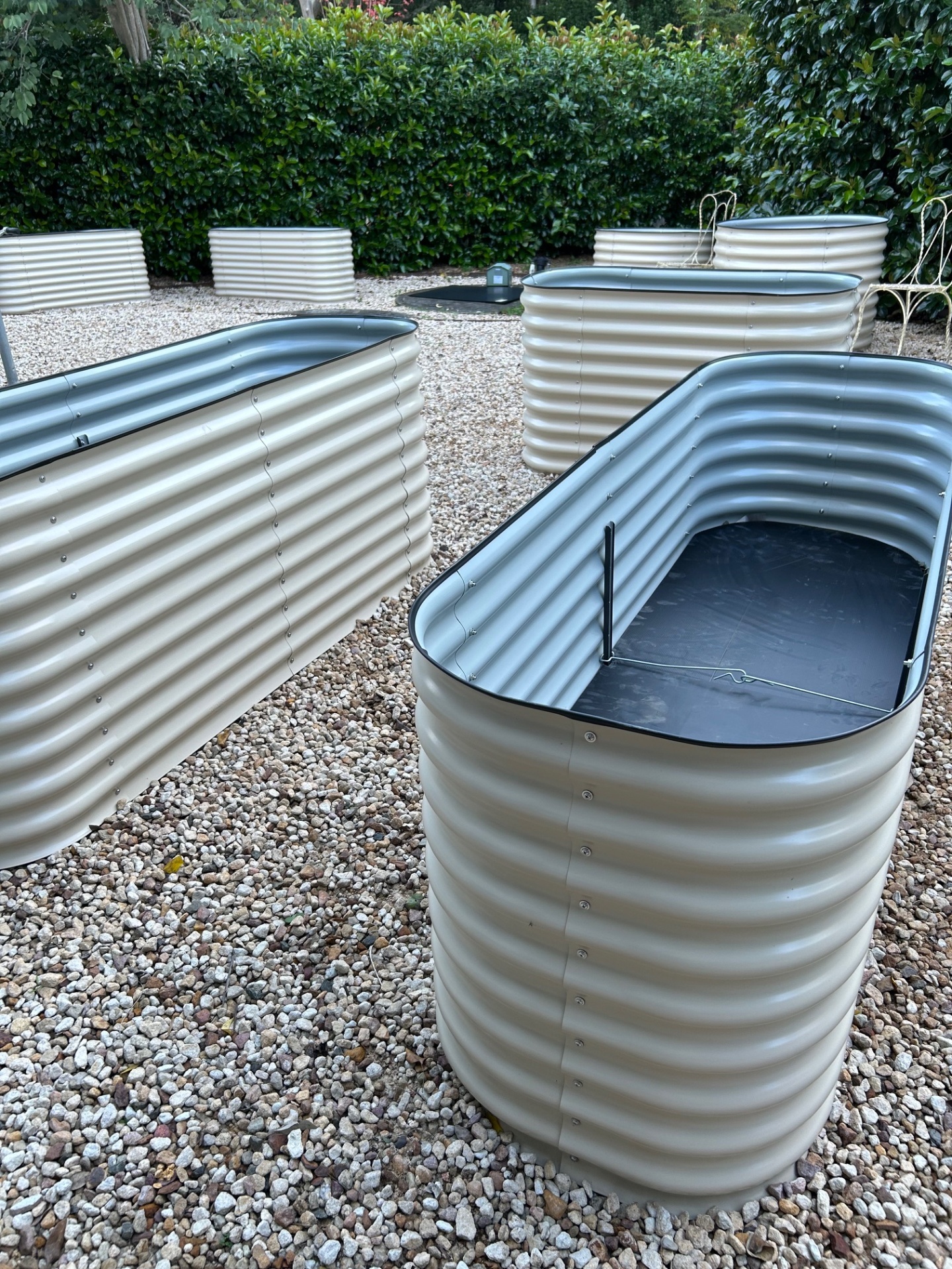 Cream coloured corrugated metal raised garden beds on gravel beside a hedge